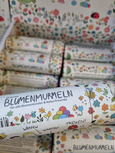 Bunte Verpackung mit Blumenmurmeln, verziert mit Naturmotiven, lädt zum spielerischen Gärtnern ein.