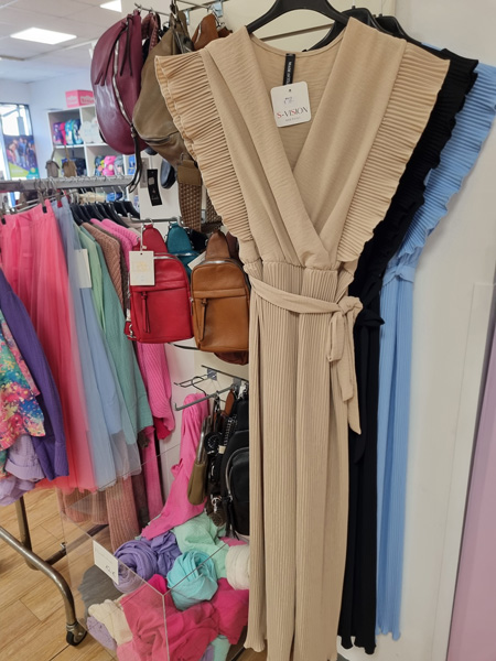 Ein beigefarbenes Plisseekleid mit Gürtel hängt in einer bunten Boutique mit Handtaschen und Kleidung.