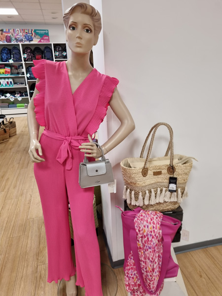 Schaufensterpuppe in pinkem Jumpsuit mit Rüschen, hält graue Handtasche. Neben ihr zwei Taschen.