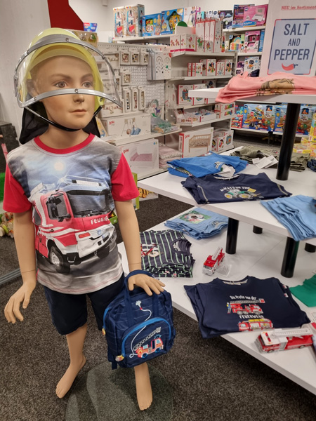 Eine Schaufensterpuppe trägt Feuerwehrkleidung für Kinder, umgeben von bunter Kinderkleidung und Spielzeug.