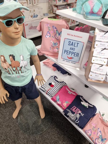 Schaufensterpuppe in mintgrünem T-Shirt und blauer Shorts, umgeben von bunten Kinder-T-Shirts im Regal.