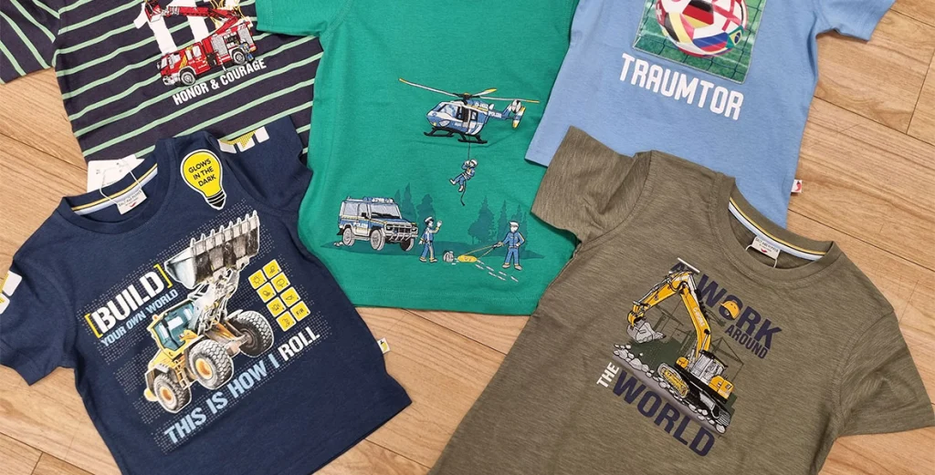 Fünf bunte Kinder-T-Shirts mit Feuerwehr-, Polizei-, Fußball- und Baggerdesigns auf Holzboden.