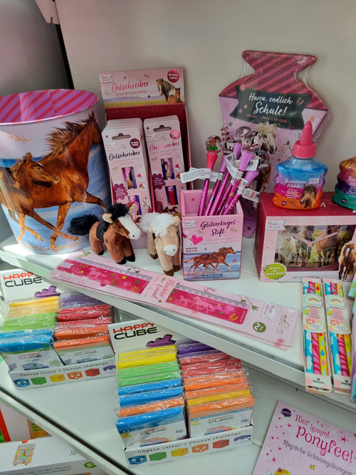 Ein Regal voller bunter Schulwaren mit Pferdemotiven, Glitzerstiften und Plüschtieren für Kinder.