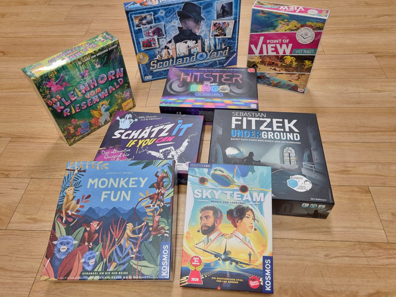 Eine bunte Sammlung von acht Brettspielen liegt auf einem Holzfußboden, bereit für einen Spieleabend.