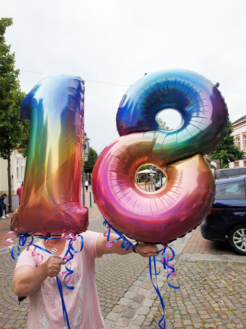 Person hält schillernde "18"-Luftballons in Regenbogenfarben, festliche Stimmung auf städtischer Straße.