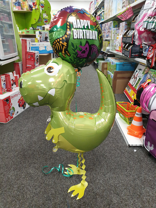 Ein grüner Dinosaurier-Ballon lächelt fröhlich unter einem bunten "Happy Birthday"-Ballon im Spielwarenladen.