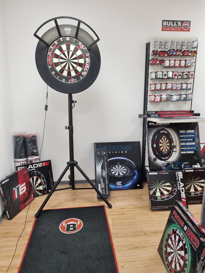 Darts-Ecke mit Dartboard, Zubehör und Verkaufsständer auf hellem Holzboden, sportliche Atmosphäre.