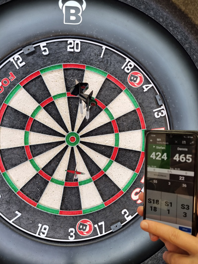 Ein Dartspiel ist im Gange; ein Smartphone zeigt den Punktestand von Stefan und führendem Dennis.