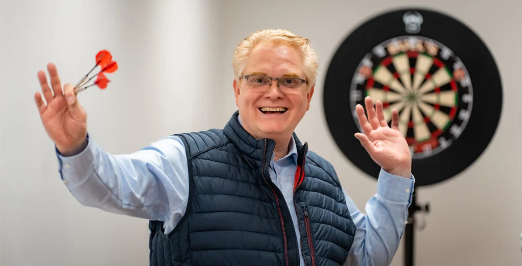 Eine fröhliche Person hält drei rote Darts vor einer Dartscheibe in einem Innenraum.